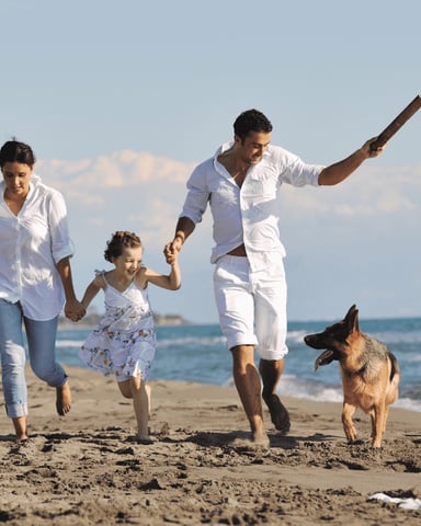 Family of four running on sandy beach with dog, ocean and waves in background