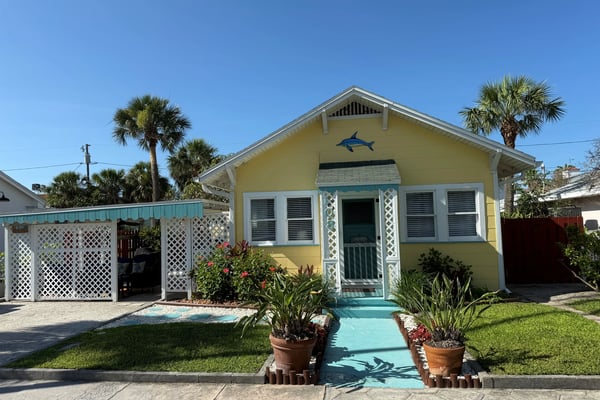 Yellow cottage with white trim and shark decoration, palm trees, lattice carport, and turquoise walkway